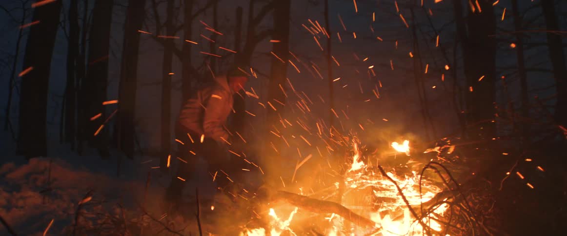 A man is putting more wood on a large bonfire in the woods on a winter evening