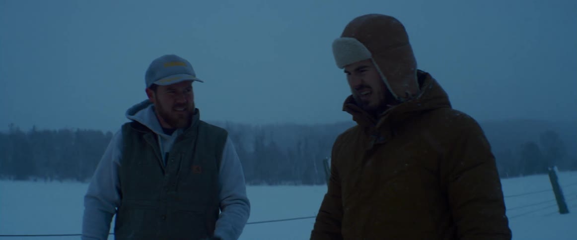 Two men in winter clothing stand and talk next to a snow covered field