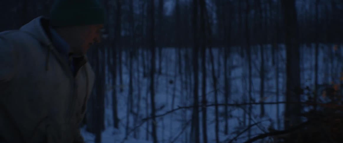 A man is building a large bonfire in the woods on a winter evening