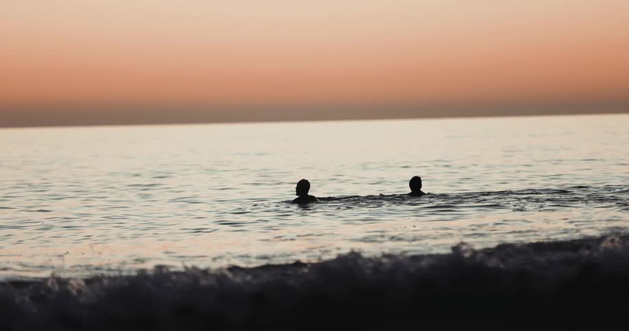 Silhouettes of two people in the water as the waves roll in under an orange sky