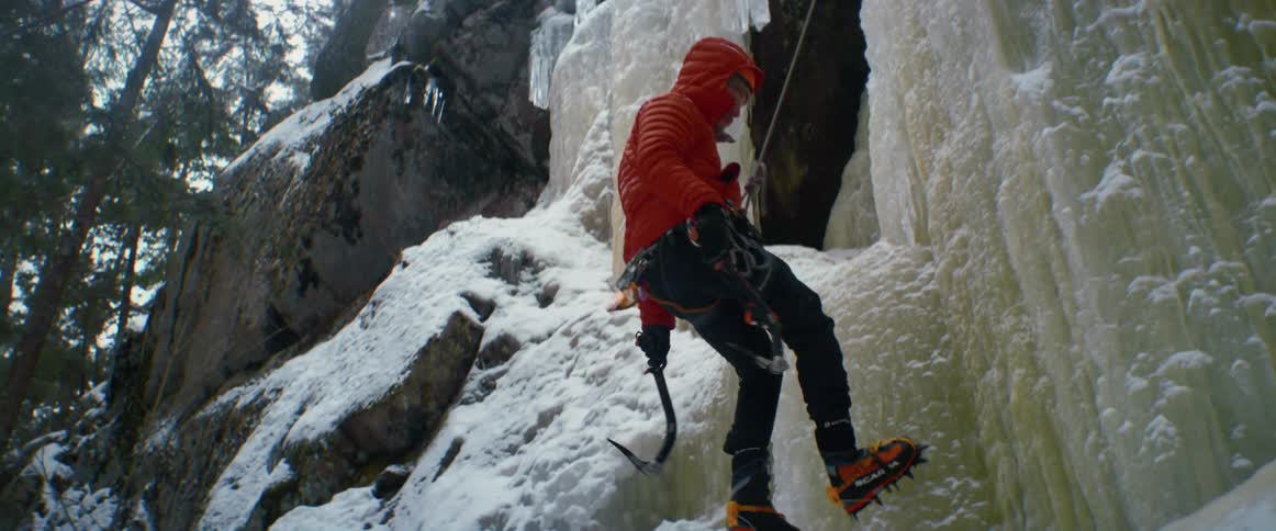 A man uses a hook and rope to climb down a frozen waterfall in a forest