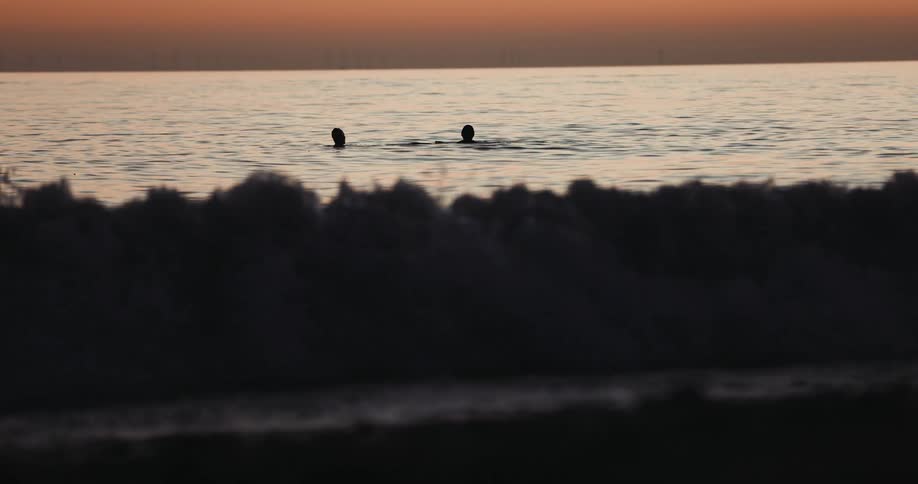 Silhouettes of two people in the water as the waves roll in under an orange sky