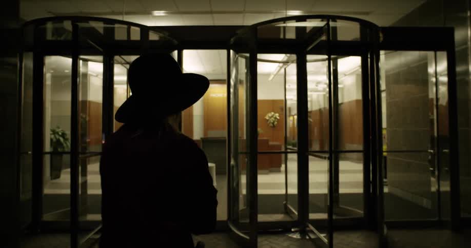 A woman in a coat and hat goes through a revolving door into a building at night