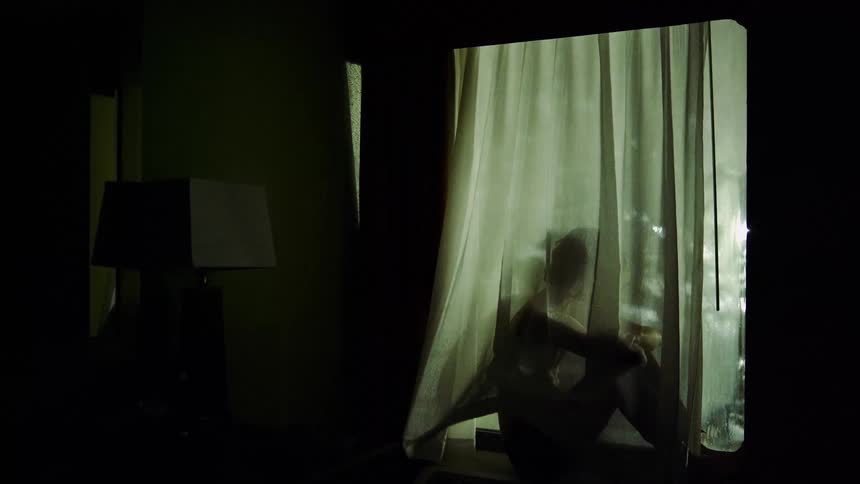 A woman sits on a window behind a curtain as she looks outside