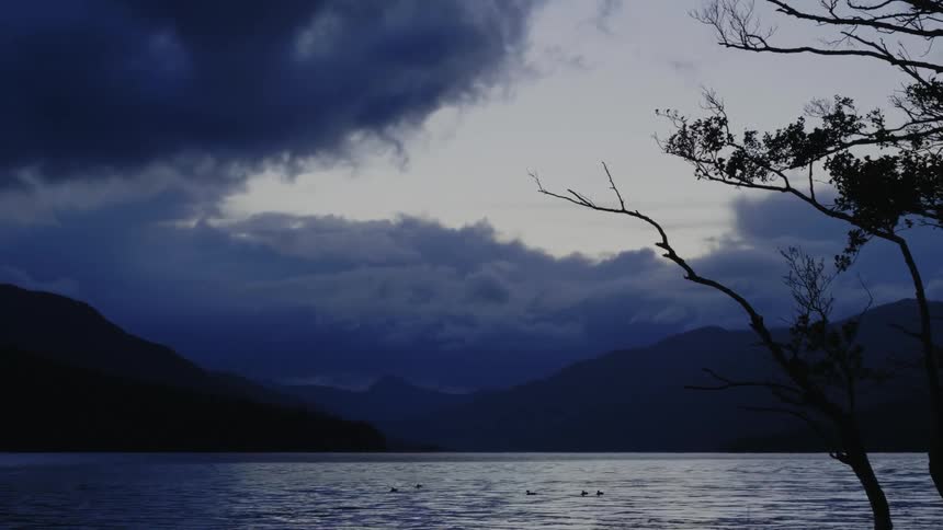 Ducks are in the water as dark clouds move overhead and there are mountains in the distance