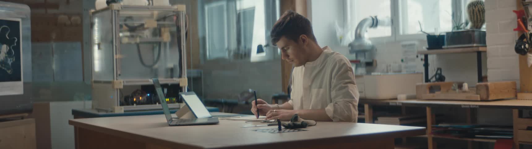 A young man is sitting at a desk with a laptop computer and a tablet and writing with a pen