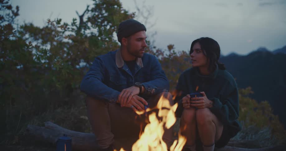 Man and woman sitting at a tree trunk at a forest area in front of a camp fire talking and holding cups