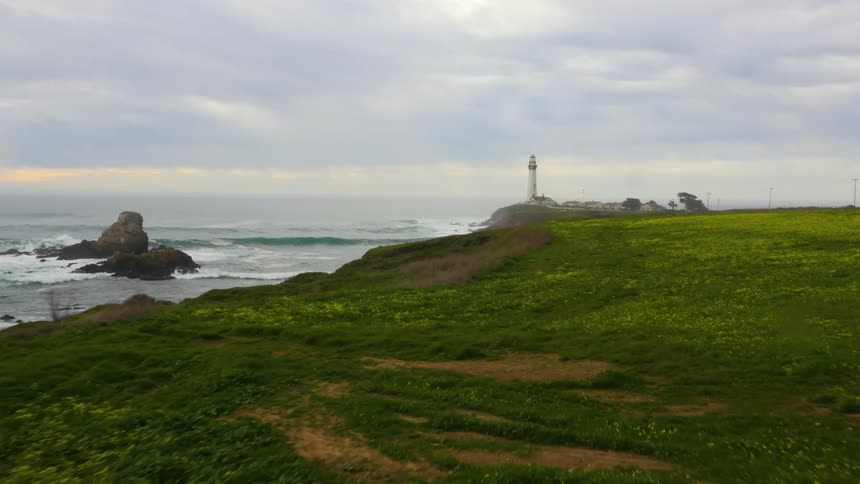 Waves splash over rocks along the coastline and a lighthouse is on a grass covered field overlooking the water