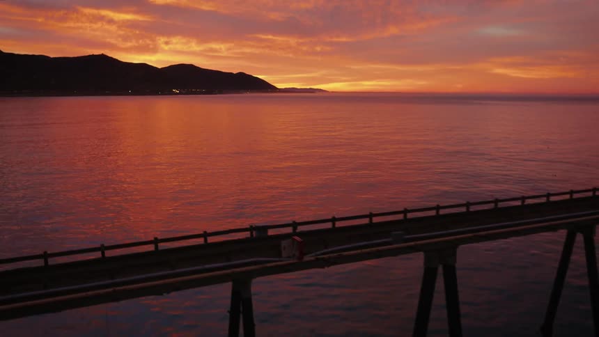 A bridge stretches across the water as it reflects the red sky and there are mountains in the distance