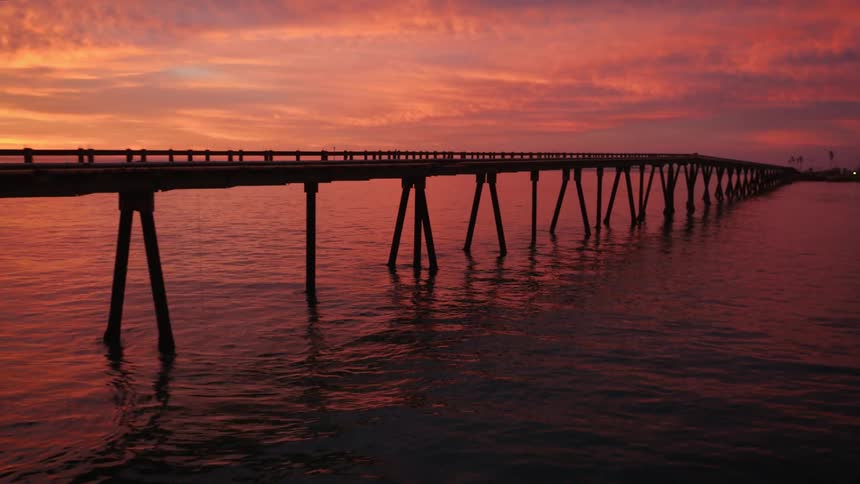 A pier stretches across the water reflecting the colors from the red sky above