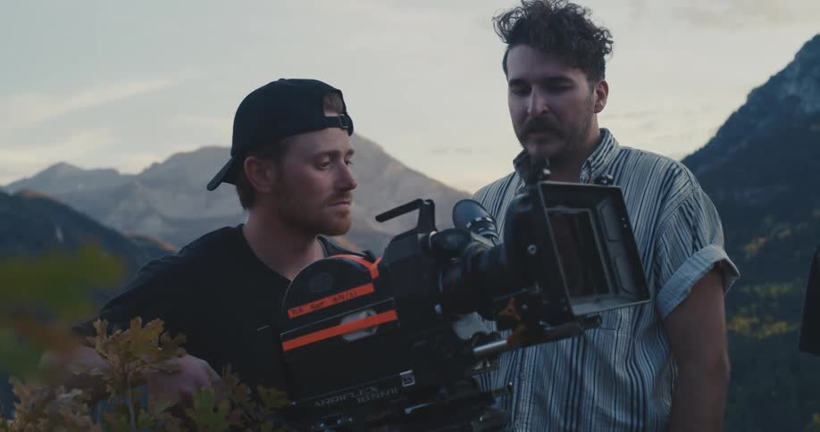 Two white men standing behind a professional cinema camera filming an outdoors shot and looking through the cameras screen