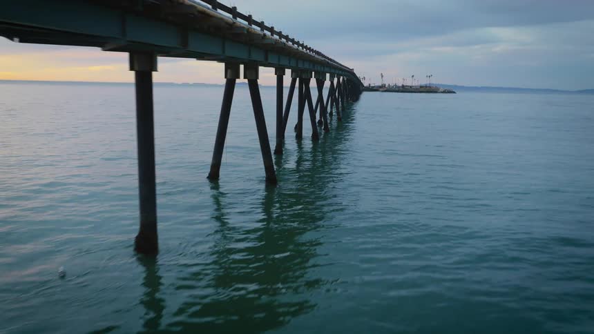 A pier stretches across the water to rocks along the shore under a cloudy sky