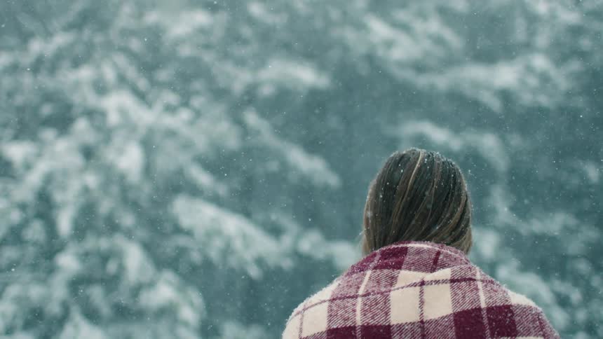 A woman walks outside as it snows with snowclad trees visible at a distance