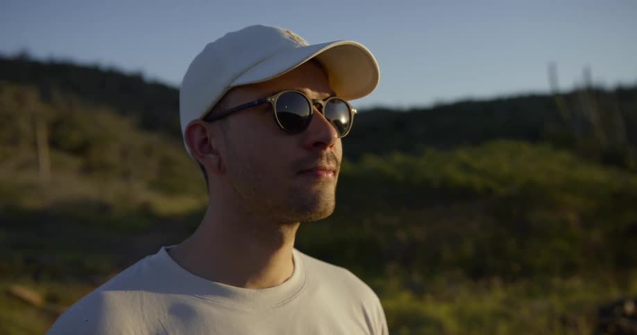 White man wearing sunglasses and a white baseball cap standing alone outdoors in the middle of a hill during day time