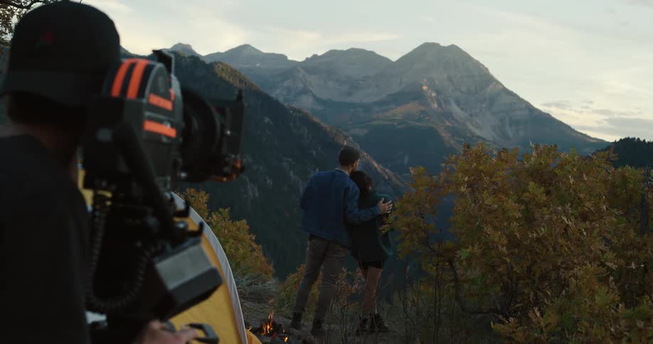 Couple hugging on the edge of a cliff near a campfire enjoying the view of mountains and nature while a cameraman films them