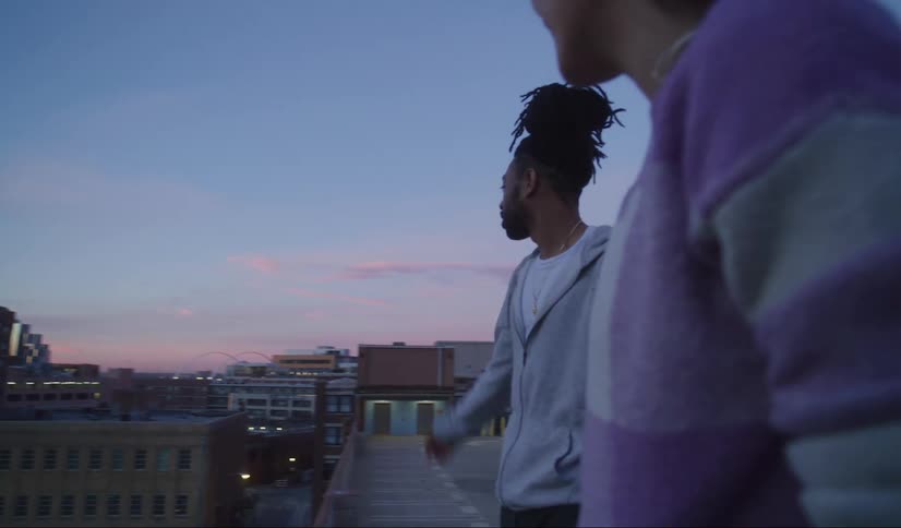 Two men are standing on a roof talking and talk as they look at the cityscape