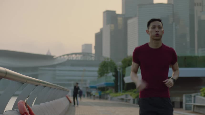Young asian man jogging at a public space in a big city with multiple tall buildings behind