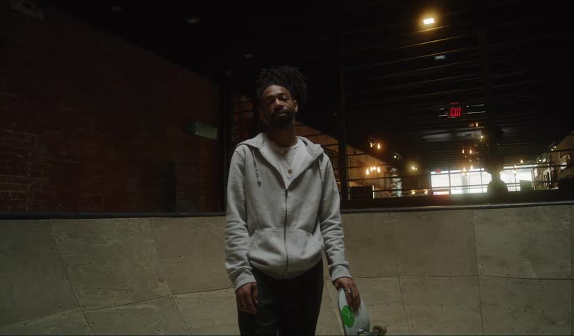 Black man wearing hoodie standing alone at an indoors skatepark holding a skateboard