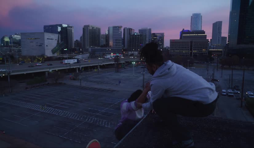 Man helps a woman up onto a rooftop as two skateboards hang over the edge