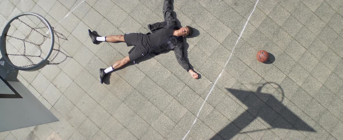 A man lies motionlessly beneath the goal on an outdoor basketball court during the day
