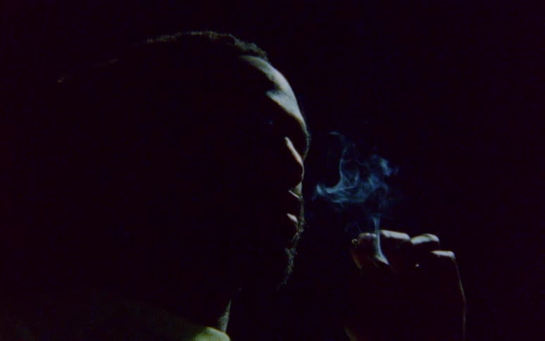 A man in a darkened room stares while holding a burning cigar