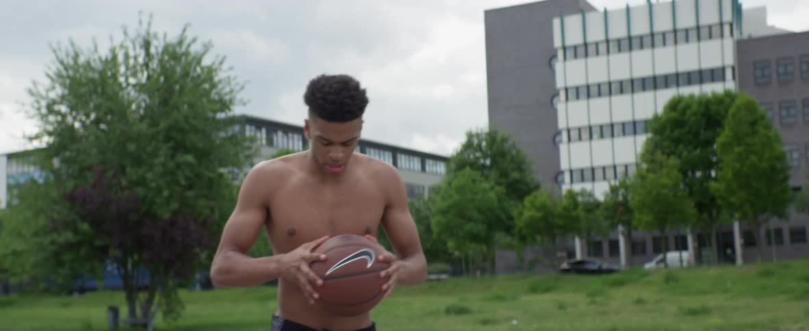 Shirtless young black man standing in the middle of a public basketball court and bouncing his basketball in the ground
