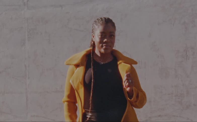 Woman with dreadlocks hoop earrings yellow overcoat and black shirt poses for the camera