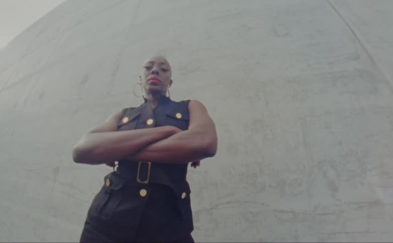 Black woman wearing hoop earrings crosses her arms as she stands in front of a cement wall