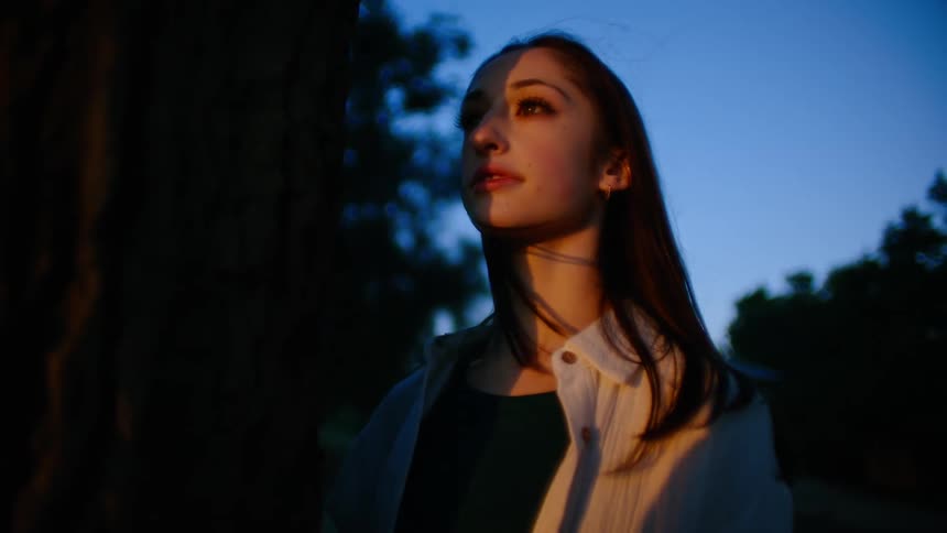 A woman is standing and staring at a sunset in a woodland area on a windy evening