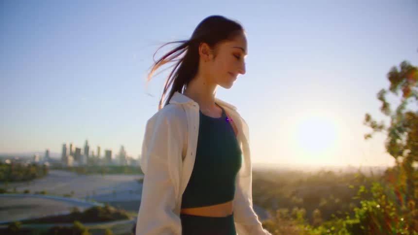 Girl with straight hair white shirt and black crop top smiles at camera standing on hill with high rise buildings in the background at sunset