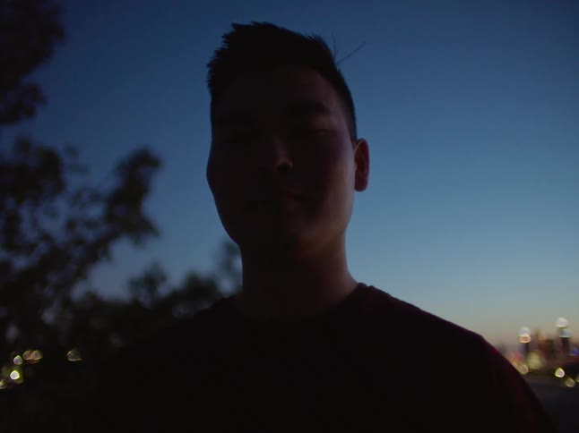 Man with black t shirt smiles at camera and is silhouetted against the sky and city skyline