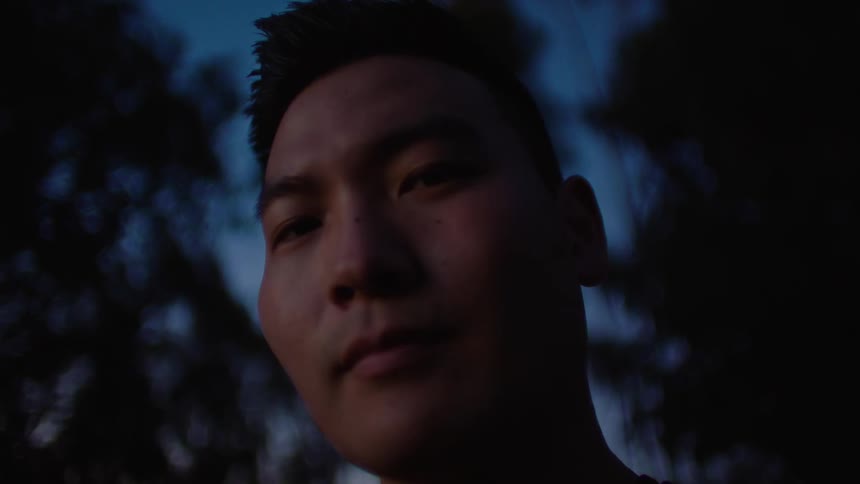 A man is staring into the camera while standing outdoors at dusk