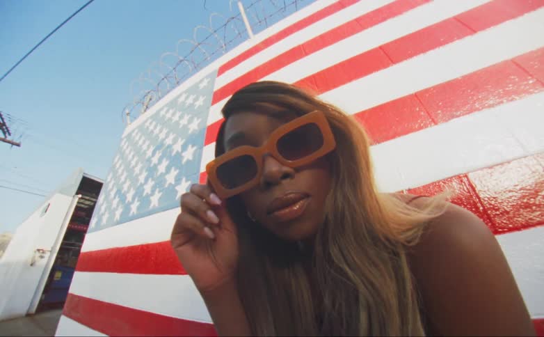 Black woman wearing orange sunglasses standing outdoors in front of american flag painted wall