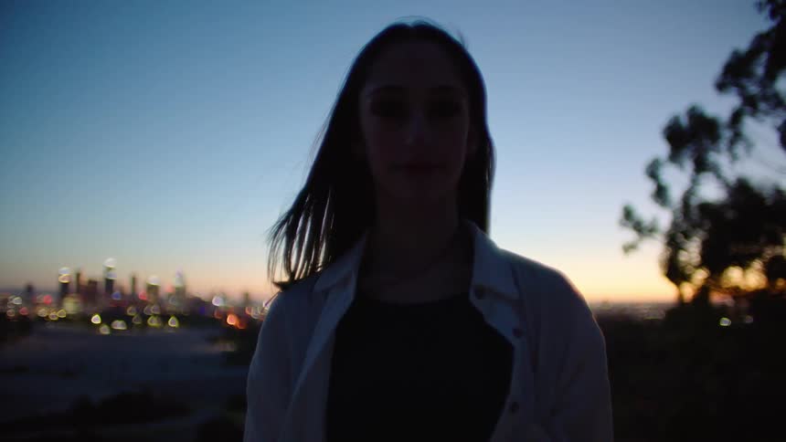 Woman with straight hair and white jacket looking at camera is silhouetted against the city skyline at dusk