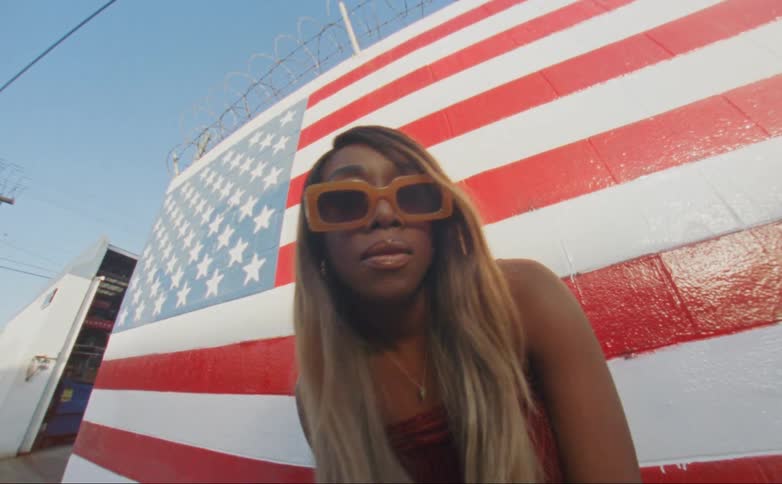A black woman posing in front of an american flag wall leans forward and tilts her head