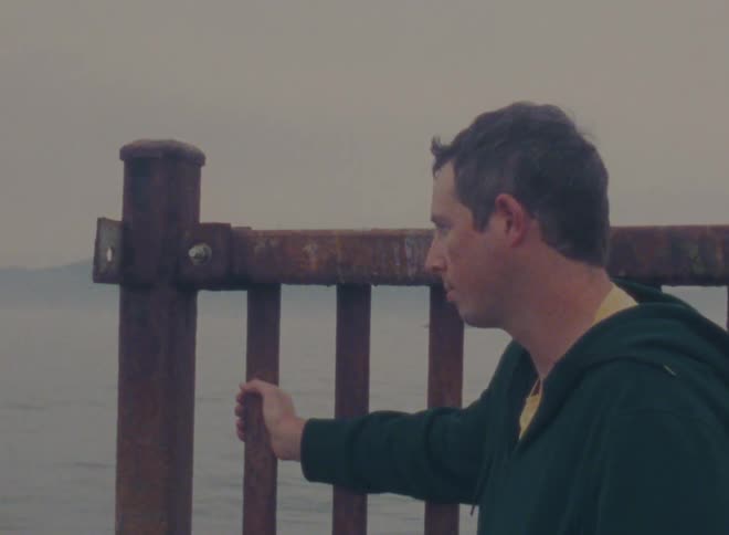 Man wearing a sweater holds onto a wooden fence as he looks out at the lake