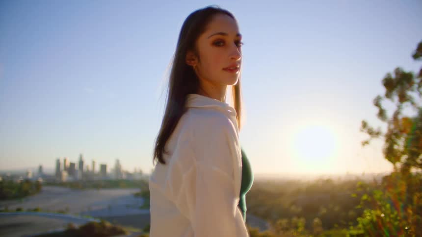 Woman with long hair stands near the trees as city buildings sit in the distance
