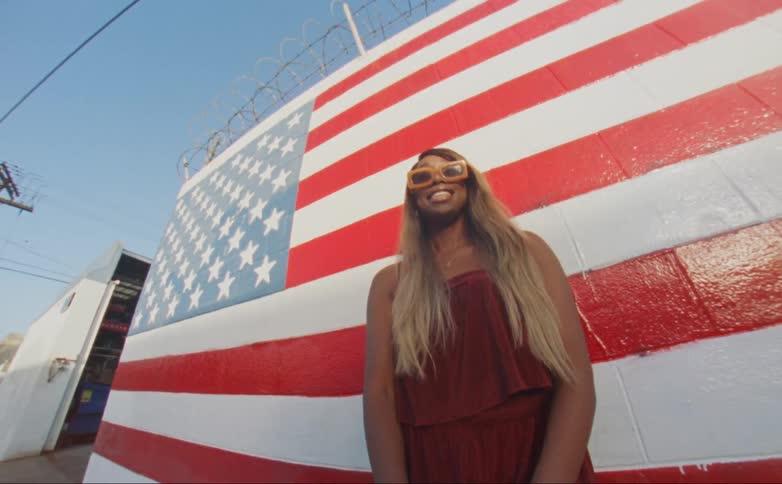 Black woman standing in front of a american flag painted wall wearing orange sunglasses and smiling