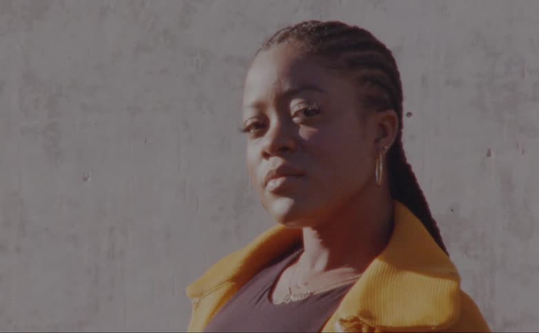 Woman with dreadlocks earrings and yellow jacket faces the camera with eye contact