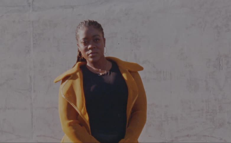 Woman with dreadlocks hoop earrings yellow overcoat and black shirt poses for the camera