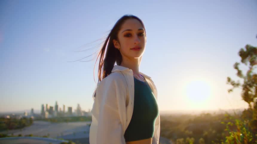 Girl with straight hair in white shirt and black cropped top smiling at camera standing on hill at sunset with tall buildings in background