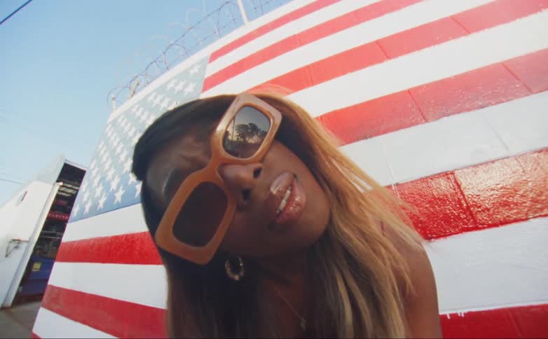 A woman leans forward and tilts her head while standing in front of a wall painted like an american flag