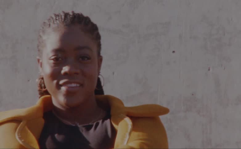 Woman with dreadlocks hoop earrings and yellow jacket faces the camera with eye contact smiling