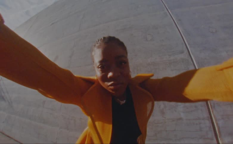 Black woman wearing a yellow overcoat dancing in front of a concrete wall outdoors