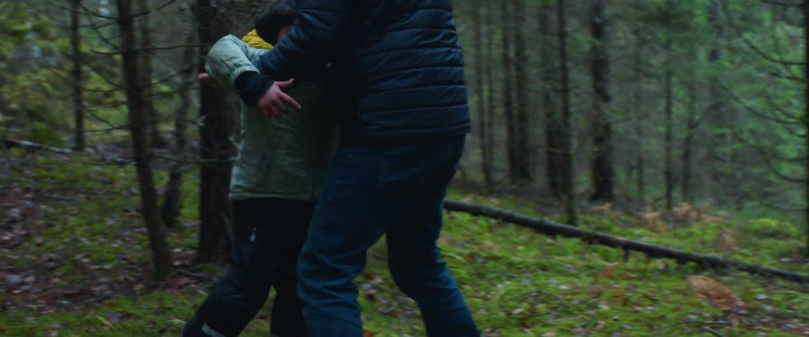 Father with navy blue coat and jeans plays with son wearing light blue coat in the forest