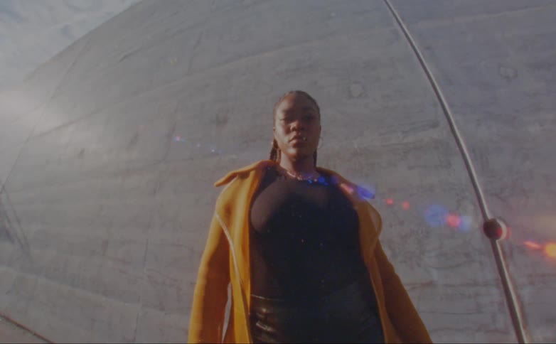 Black woman wearing a yellow overcoat posing for the camera in front of a concrete wall outdoors at a sunny day