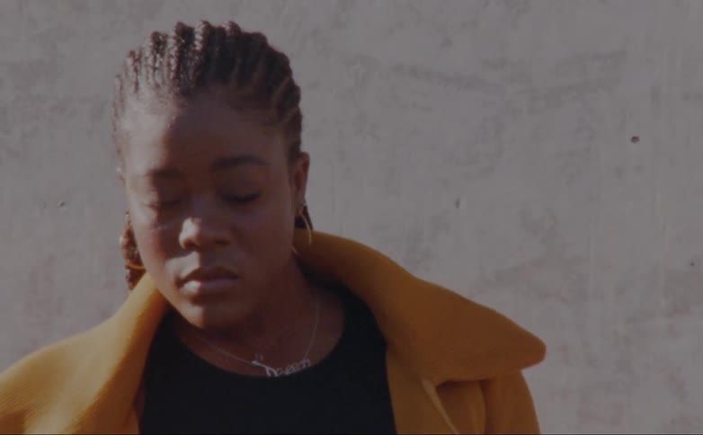 Black woman standing in front of a concrete wall wearing a yellow overcoat and posing for the camera