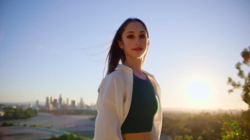 Girl with straight hair white shirt and black cropped top smiling at camera standing on hill at sunset with tall buildings in background