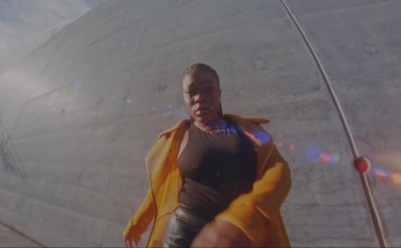 Black woman wearing a yellow overcoat dancing in front of a grey concrete wall during day time