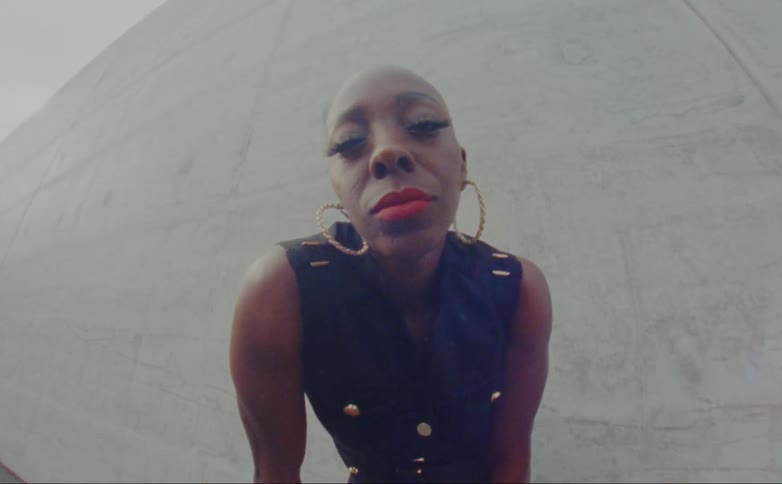 Bald female model with hoop earrings and black dress looks down at camera